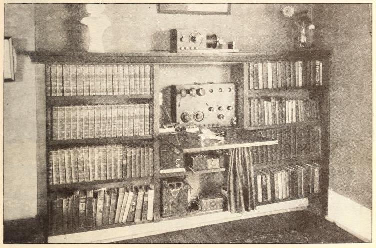 A scan of a printed black-and-white photograph. The photo shows a bookshelf filled with large books. The center shelf contains a radio set and a fold-out table.