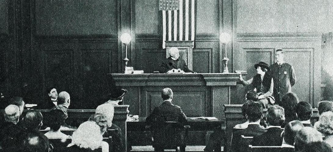 A black and white photograph of a courtroom. A judge in robes is sitting at and elevated desk. There is a witness beside them, and an attorney facing them. There are many people in the crowd