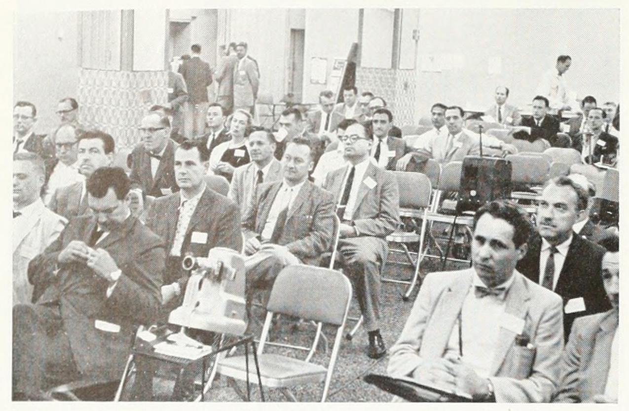 A black and white photo of several people sitting in rows of chairs and watching a presentation.